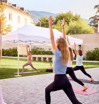 The white folding gazebo is in use at a yoga class. The yoga instructor is standing under it. In front of the gazebo are the participants in the yoga class.