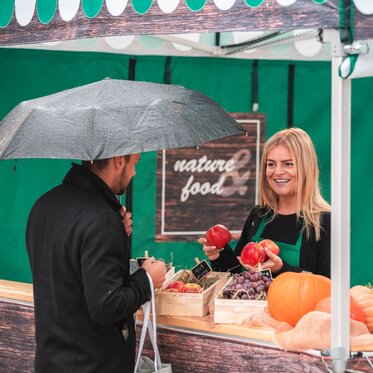 La venditrice nella tenda del mercato tiene in mano diversi frutti. Il cliente è in piedi davanti alla tenda di vendita al banco.