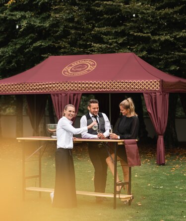 Eleganter Faltpavillon 8x4 m in Burgunderrot mit goldenen Verzierungen und dekorativen Vorhängen, die eine Stange bedecken. Der Pavillon steht auf einer Wiese und wird für den Empfang einer eleganten Veranstaltung verwendet, während die Leute Sekt von einer Kellnerin serviert trinken