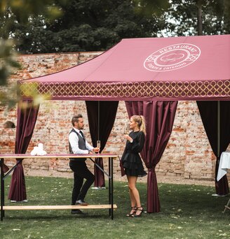 Il gazebo catering, di colore bordeaux, si trova nell'ampio giardino. La donna e l'uomo hanno un bicchiere di champagne in mano e sono in piedi davanti ad un tavolo alto.