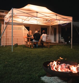 Das 4,5x3 m Gartenzelt steht im Dunkeln im Garten. Darunter sitzen die 4 Gäste. Vor dem Zelt ist noch eine Feuerstelle.