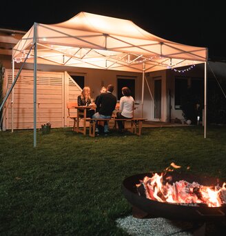 Il gazebo da giardino di 4,5x3 m si trova in giardino al buio. I 4 ospiti siedono sotto di essa. Davanti al gazebo c'è un camino.