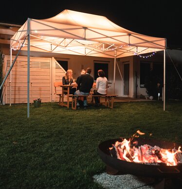 The 4.5x3 m garden tent stands in the garden in the dark. The 4 guests sit under it. There is a fireplace in front of the gazebo.