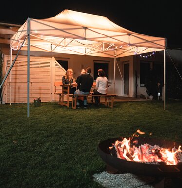 Il gazebo da giardino di 4,5x3 m si trova in giardino al buio. I 4 ospiti siedono sotto di essa. Davanti al gazebo c'è un camino.