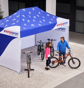 Das Promotionzelt von Intersport ist ein 4,5x3 m großer Faltpavillon. Er ist vollflächig bedruckt in weiß-blau. Auf dem Dach sind verschiedene Sport-Icons zu sehen. Der Verkäufer zeigt der Kundin das Fahrrad.