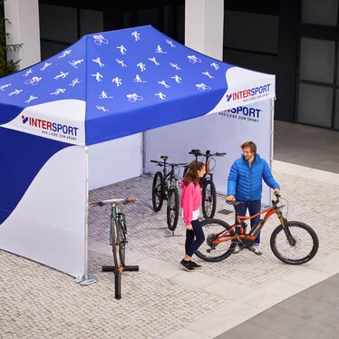 Das Promotionzelt von Intersport ist ein 4,5x3 m großer Faltpavillon. Er ist vollflächig bedruckt in weiß-blau. Auf dem Dach sind verschiedene Sport-Icons zu sehen. Der Verkäufer zeigt der Kundin das Fahrrad.