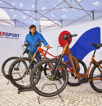 Il venditore si trova sotto il gazebo pieghevole. Sotto ci sono anche delle biciclette. Il gazebo è stampato nel corporate design di Intersport.