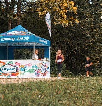 Das blau-weiße Eventzelt dient als Versorgungsstation bei einem Marathon. 2 Läufer laufen gerade am Eventzelt vorbei.