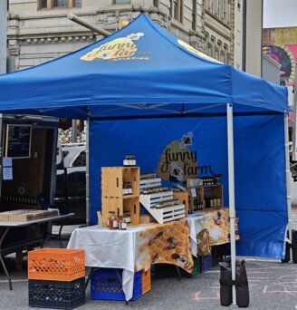 Auf einem Markt steht ein blauer, bedruckter Ecotent Faltpavillon Serie E1 der Größe 3 x 3 Meter mit Seitenwand von Funny Farm. 