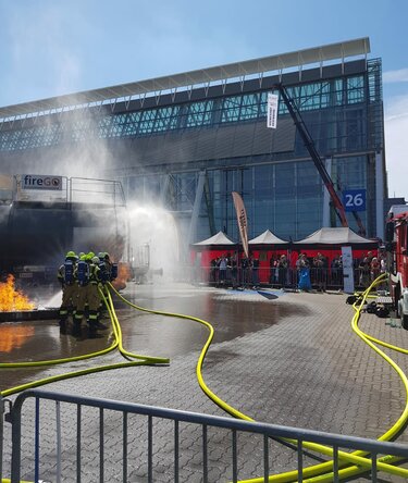 Squadra di vigili del fuoco durante una dimostrazione antincendio all’aperto in fiera, con getto d’acqua, veicoli di soccorso e gazebo di emergenza sullo sfondo.